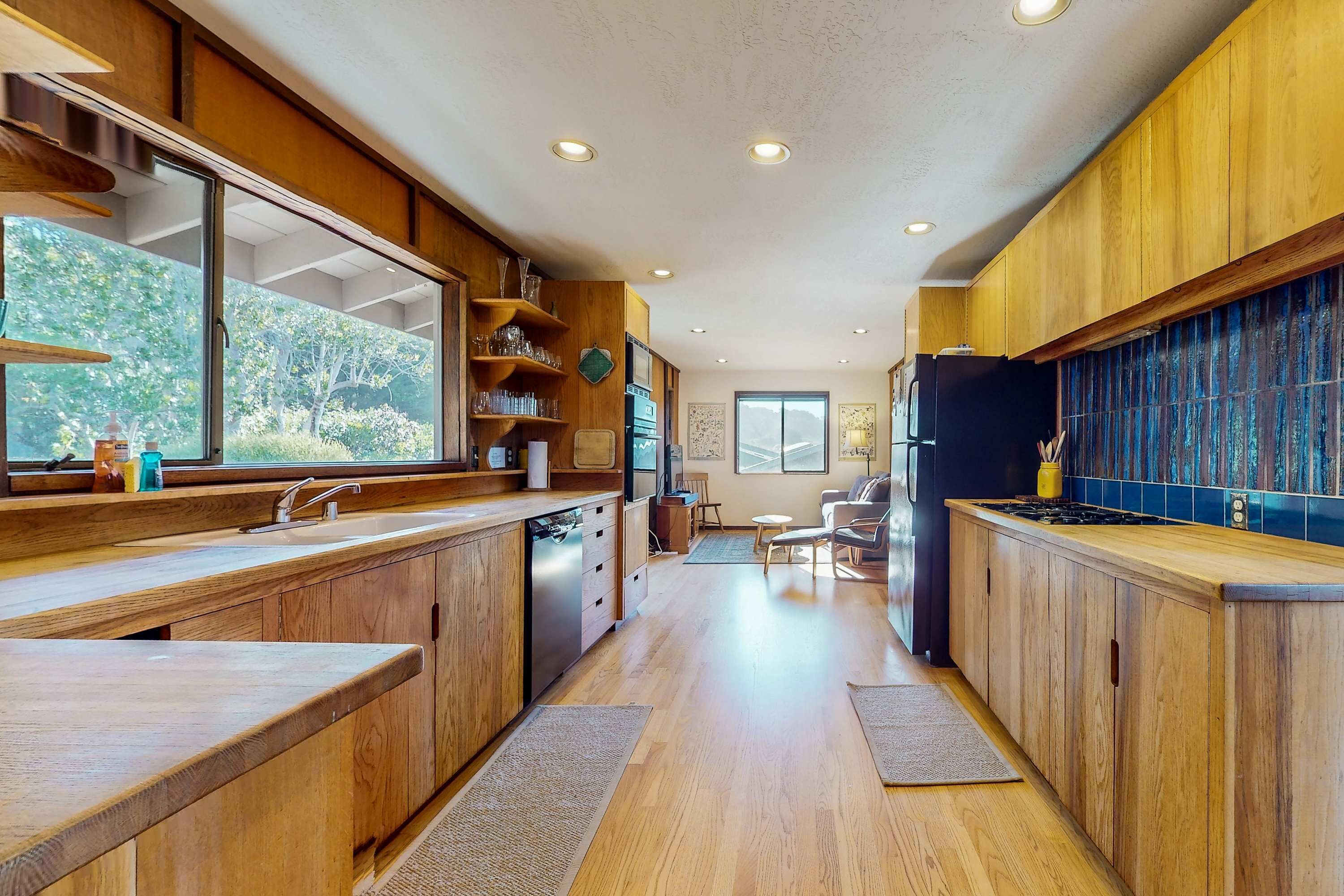 A modern kitchen with wooden cabinets, a built-in oven, and a large window overlooking a forested area.