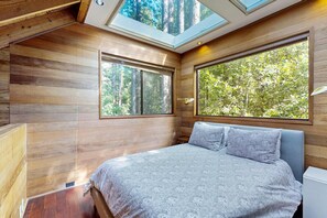 A bedroom with a bed, skylights, and large windows with a forest view. - Forested cabin with private hot tub & ocean views (Sea Ranch)