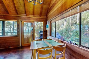 A wooden interior with a dining area, large windows, and a view of trees. - Forested cabin with private hot tub & ocean views (Sea Ranch)