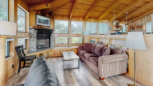 Een gezellig chaletinterieur met een stenen open haard, houten balken, een bank, een tafel en een lamp.