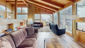 Een ruime woonkamer met een houten plafond, grote ramen, een bruine bank, een zwarte fauteuil en een houten salontafel.