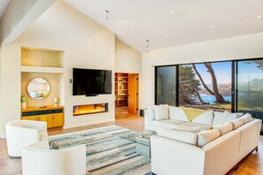 A modern living room with a large TV, white sofas, a fireplace, and a large window with a coastal view. - Oceanfront home with fireplace, EV charger & ocean views (Sea Ranch)