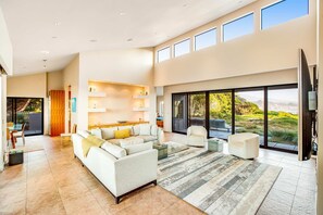 A spacious living room with a large sectional sofa, a coffee table, and a TV. There are multiple windows providing a view of the outdoors. - Oceanfront home with fireplace, EV charger & ocean views (Sea Ranch)