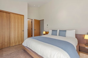 A hotel room with a large bed, two bedside lamps, a wooden wardrobe, and a door leading to another room. - Oceanfront home with fireplace, EV charger & ocean views (Sea Ranch)
