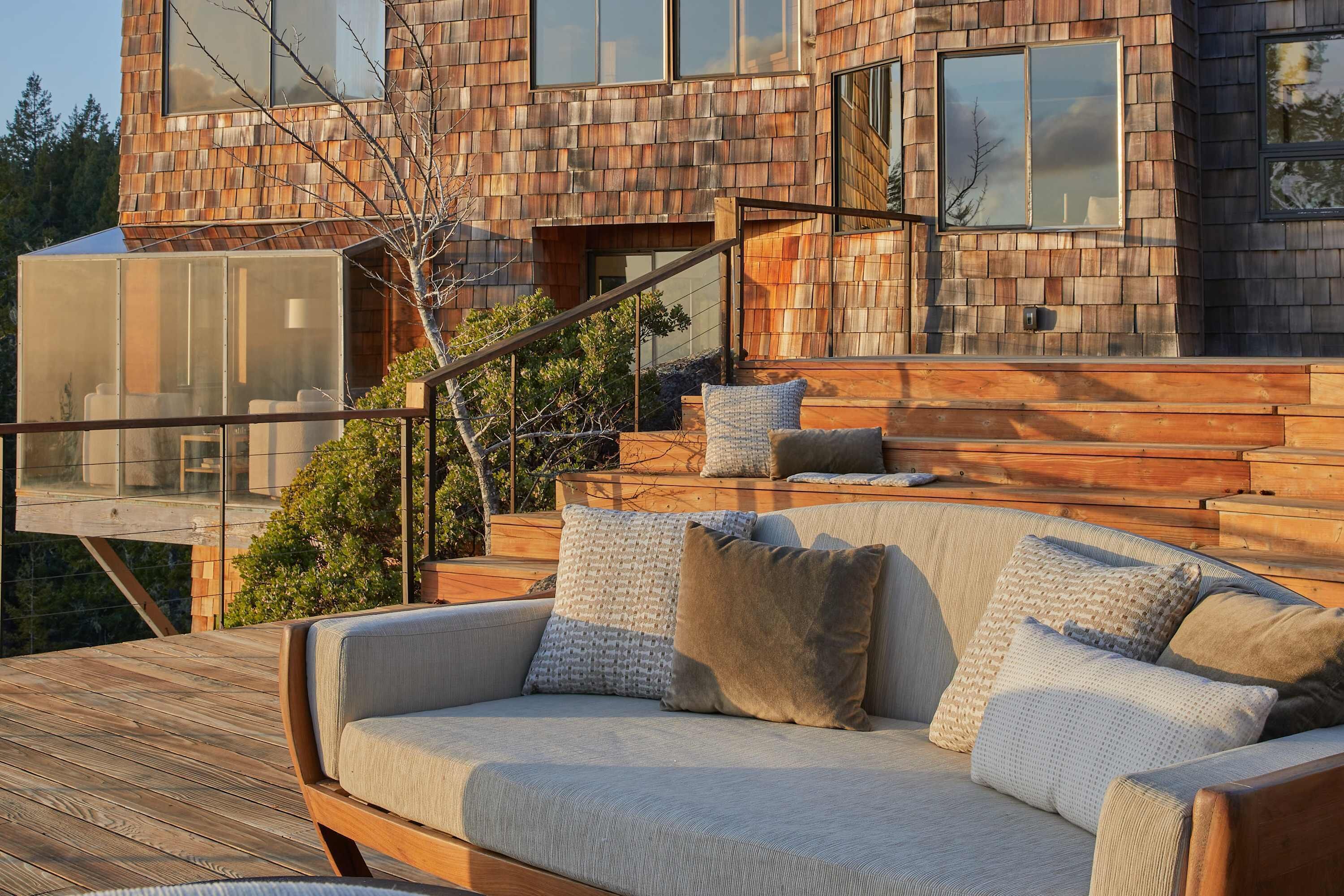 Une maison moderne avec une façade en bois, une véranda en verre et une terrasse en bois agrémentée d’un canapé et de coussins.