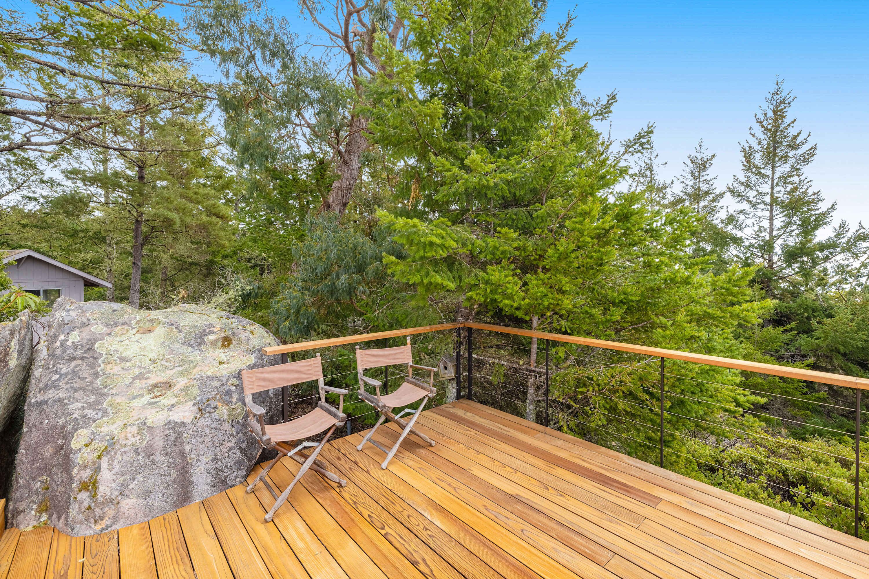 Una terraza de madera con sillas, rodeada de árboles y una roca grande.