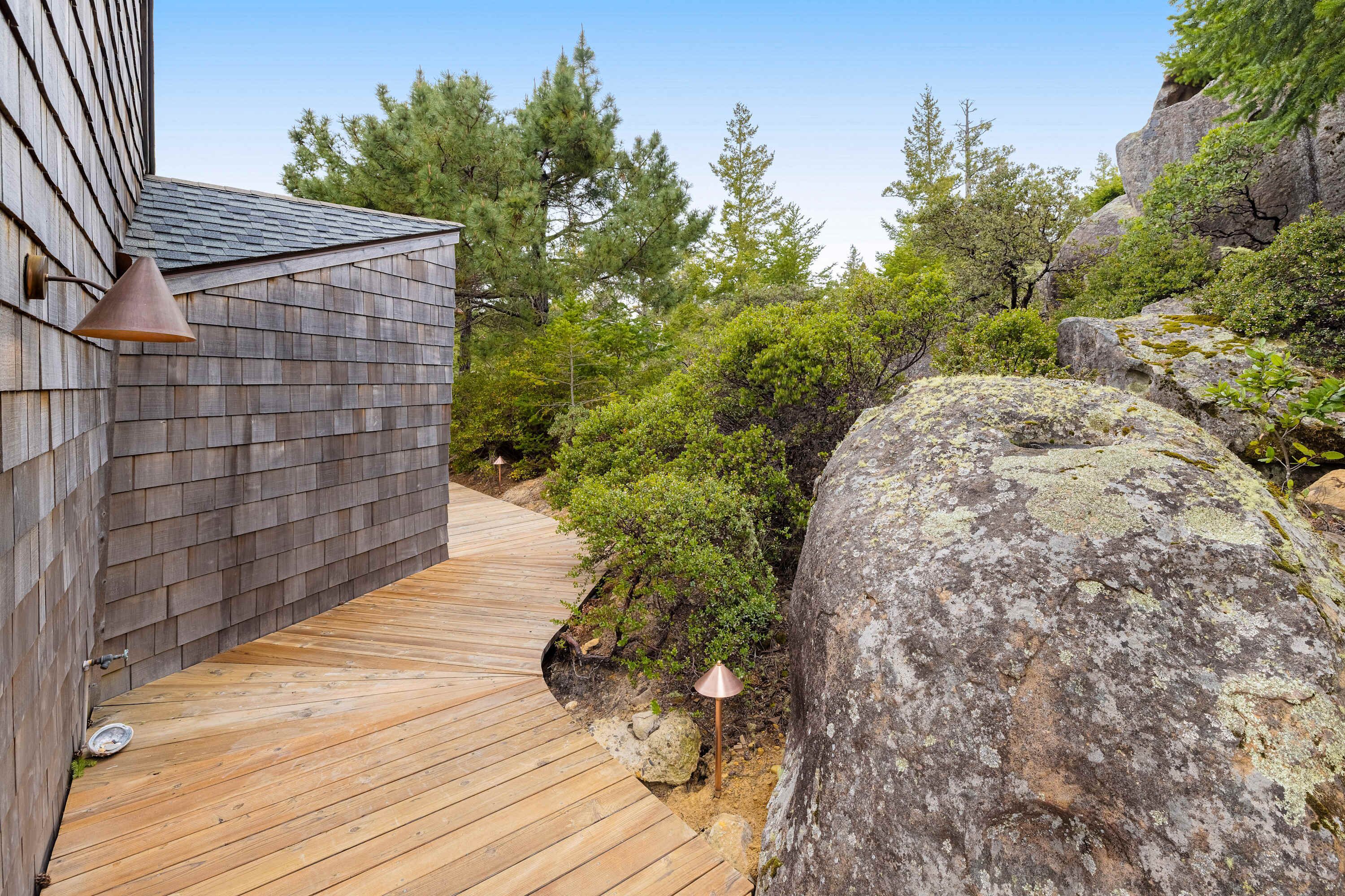A wooden deck leading to a building with a sloped roof, surrounded by dense greenery and large rocks.