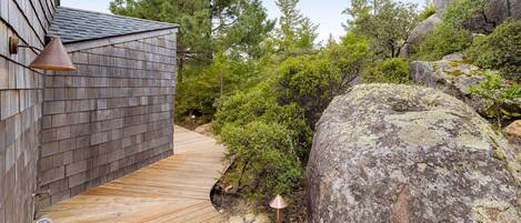 A wooden deck leading to a building with a sloped roof, surrounded by dense greenery and large rocks.