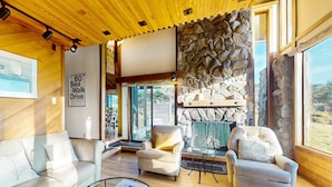 Un amplio salón con chimenea de piedra, techo de madera y grandes ventanales que ofrecen vistas al exterior.