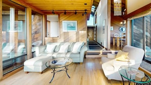 Una sala de estar moderna con techo de madera, paredes de vidrio, un sofá, una mesa de centro de vidrio y una escalera que conduce a otro nivel.
