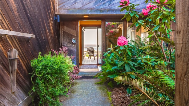 Fachada de madera con puerta de cristal, pequeño sendero y abundante vegetación, incluyendo flores rosadas y helechos.