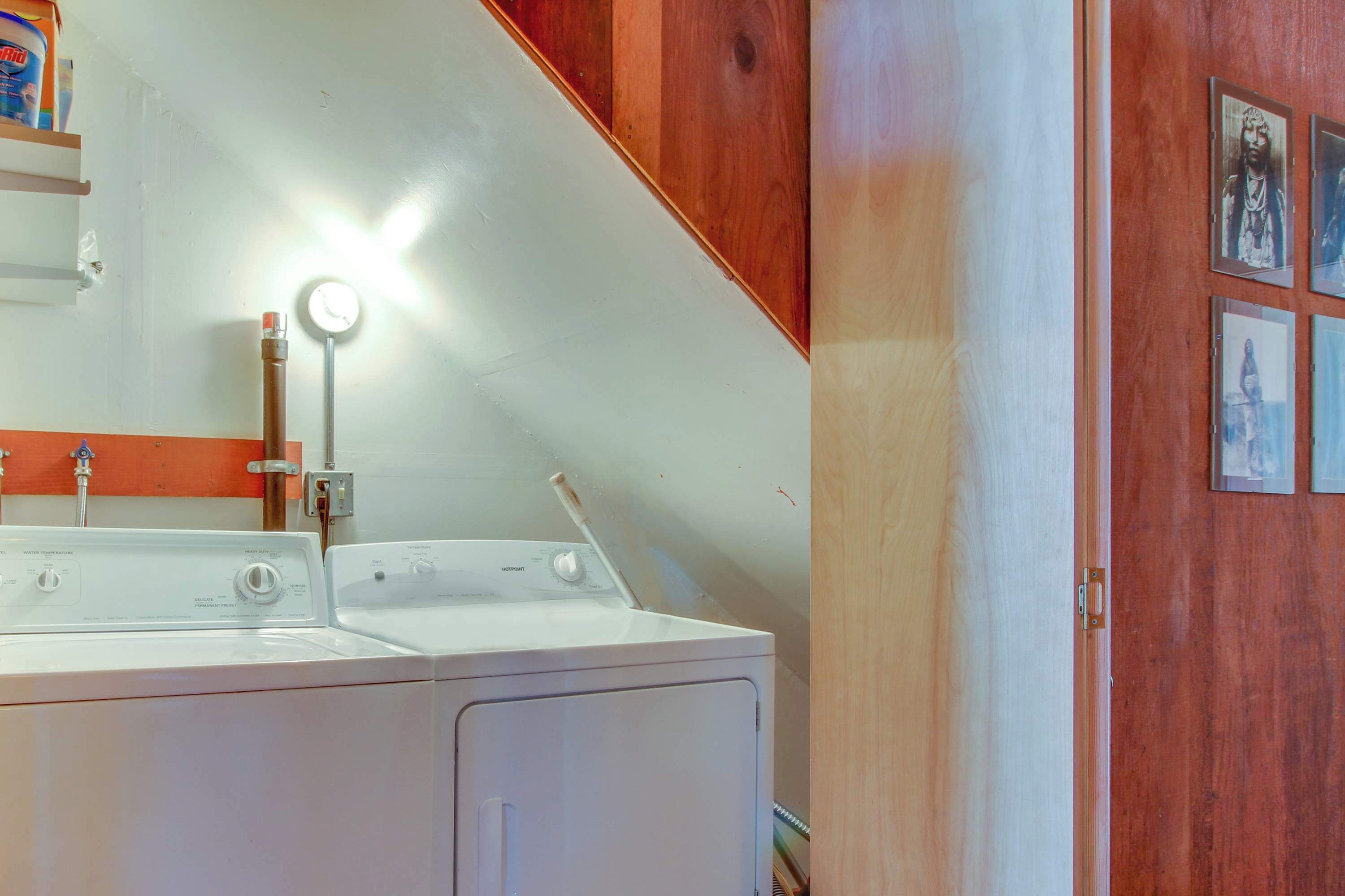 A laundry area with two white washing machines, a wooden shelf with a detergent box, and a wall-mounted light fixture.