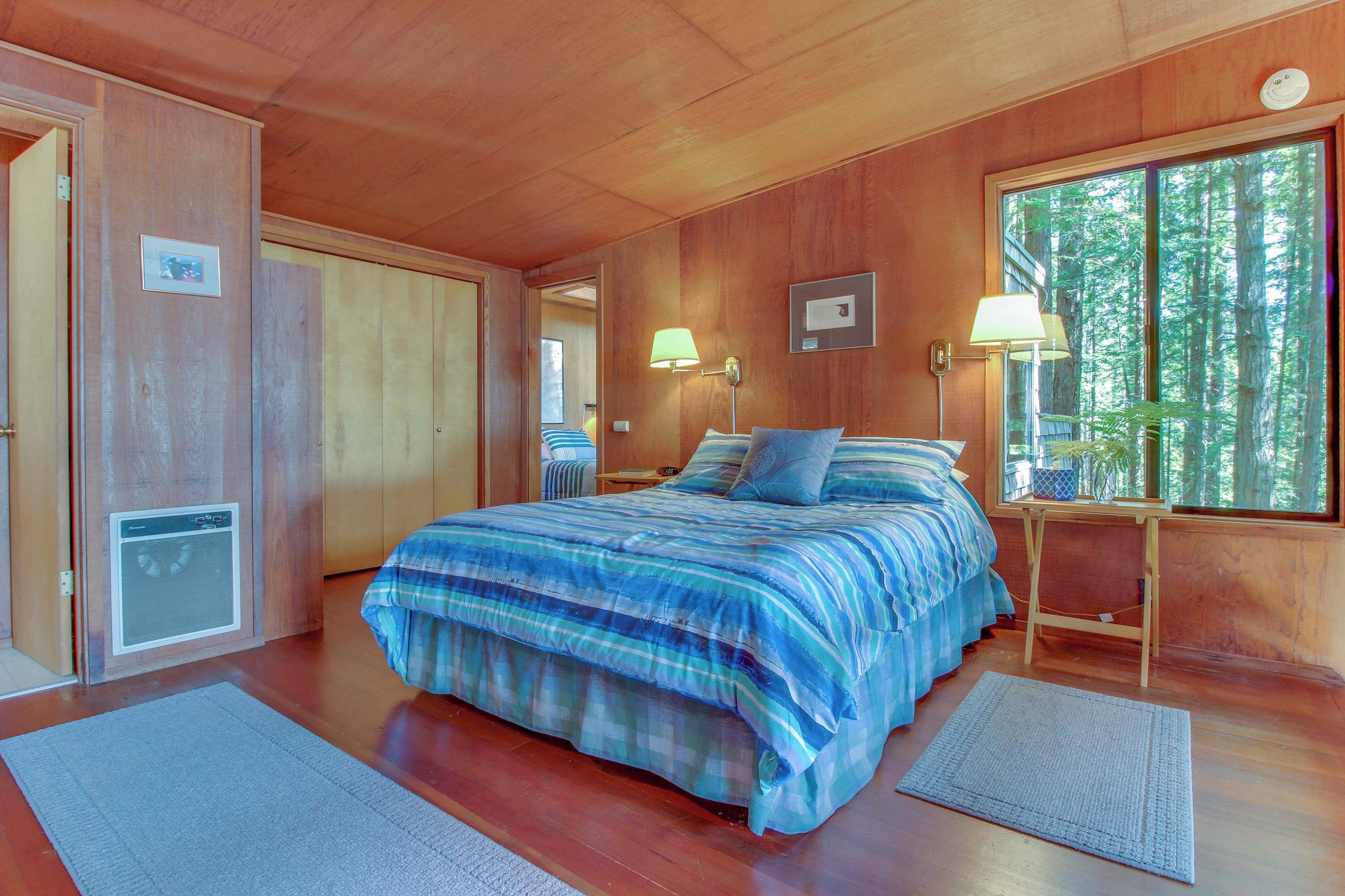 Un dormitorio con cama, dos lámparas de noche, ventana con vista a árboles y techo de madera.