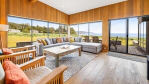 Ein modernes Wohnzimmer mit einem Ecksofa, Holzboden, großen Fenstern und Blick ins Freie.