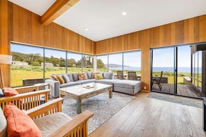 Ein modernes Wohnzimmer mit einem Ecksofa, Holzboden, großen Fenstern und Blick ins Freie.
