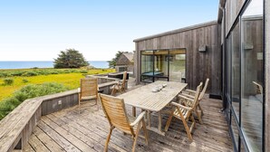 Eine Holzterrasse mit Tisch und Stühlen, mit Blick auf eine Wiese und das Meer.