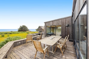 Eine Holzterrasse mit Tisch und Stühlen, mit Blick auf eine Wiese und das Meer.