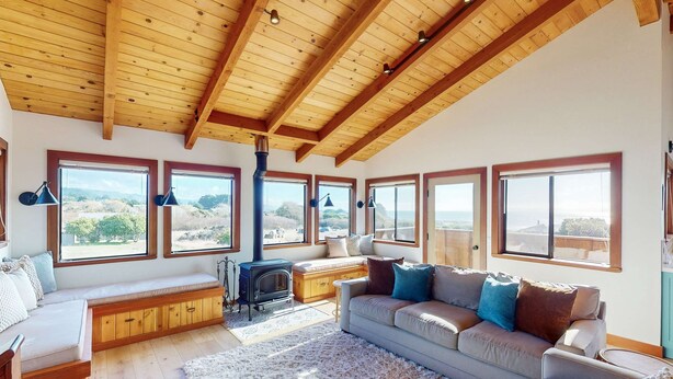 Uma sala de estar ampla com teto de madeira alto, assentos embutidos, lareira e grandes janelas com vista para o exterior.