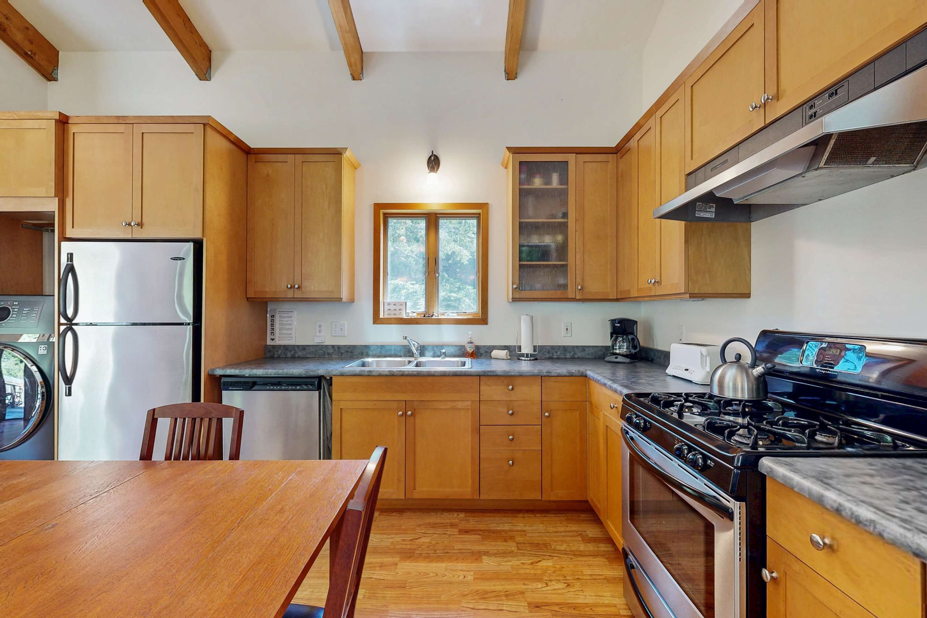 A kitchen with wooden cabinets, a stainless steel refrigerator, a stove, and a window with a view of trees.