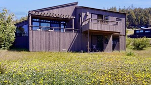 Una casa moderna de madera con amplios ventanales, un balcón y vistas a un campo lleno de flores amarillas.