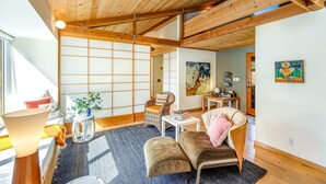 En rummelig opholdsstue med højt til loftet, træbjælker, tatami-måtter, en sofa, en lænestol, et lille bord og en reol.