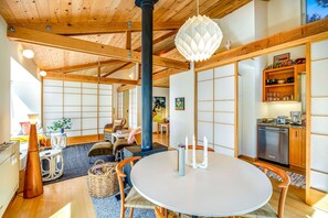 Ein traditioneller japanischer Raum mit Tatami-Böden, einem Tisch aus Tatami-Matten, einer schwarzen Säule, einer dekorativen Pendelleuchte und einer Küchenzeile mit Holzschränken und einem Kühlschrank.