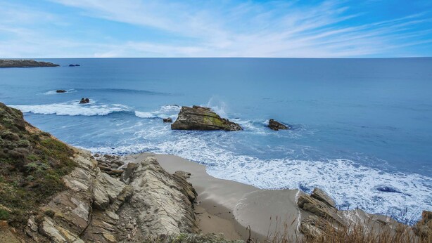 Paisagem costeira com falésias rochosas, uma praia arenosa e o oceano.