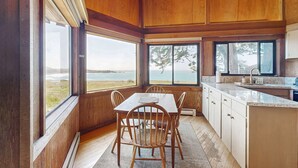 Un intérieur en bois comprenant une table et des chaises, un coin cuisine et de grandes fenêtres donnant sur l’océan.