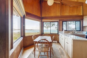A wooden interior with a dining table and chairs, a kitchen area, and large windows offering a view of the ocean. - Oceanfront home with fireplace near Black Point Beach, dogs welcome (Sea Ranch)