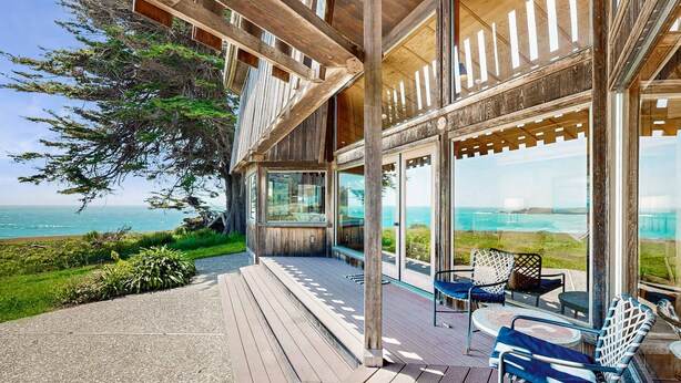 Une terrasse en bois avec des sièges d’extérieur, de grandes fenêtres et une vue sur l’océan.