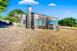 A modern wooden house with large windows, a chimney, and a fenced yard. - Ocean-view meadow home with deck & grill near Black Point, dogs welcome (Sea Ranch)