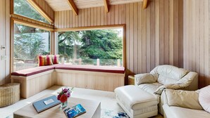 Une pièce avec un plafond en bois, une grande fenêtre donnant sur les arbres, un canapé blanc et une petite table sur laquelle se trouvent un livre et des fleurs.