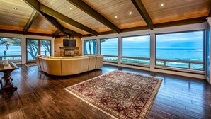 Une pièce spacieuse avec un grand plafond en bois, un canapé beige, un tapis à motifs et une vue sur l’océan par de grandes fenêtres.