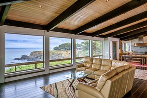 A spacious living area with a leather sofa, a glass coffee table, and large windows offering a view of the ocean and cliffs. - Blufftop home with hot tub & forever oceanfront views & private cottage (Gualala)