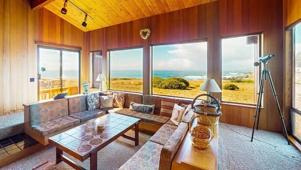 A room with a wooden ceiling, large windows with a beach view, a sofa, a table, and a tripod. - Sea Ranch 2BR getaway with ocean views, wood stove & private deck (Sea Ranch)