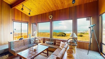 A room with a wooden ceiling, large windows with a beach view, a sofa, a table, and a tripod.