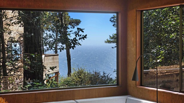 Een kamer met een groot raam, met uitzicht op zee en bomen.