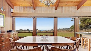A wooden dining table with chairs, a chandelier, and large windows with a view of the landscape. - Sea Ranch 3BR distant ocean-view home with hot tub — dogs welcome (Sea Ranch)