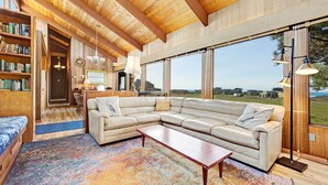 A spacious living area with a large sectional sofa, a coffee table, and a colorful rug. There are bookshelves and a dining area visible in the background. - Sea Ranch 3BR distant ocean-view home with hot tub — dogs welcome (Sea Ranch)