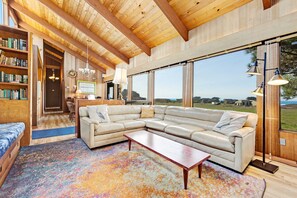 A spacious living area with a large sectional sofa, a coffee table, and a colorful rug. There are bookshelves and a dining area visible in the background. - Distant ocean-view home with hot tub near Walk On Beach, dogs welcome (Sea Ranch)