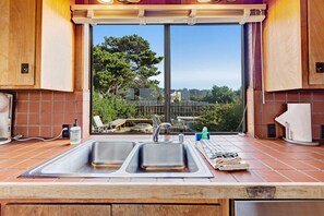 A kitchen with a large window, two sinks, and a view of greenery outside. - Distant ocean-view home with hot tub near Walk On Beach, dogs welcome (Sea Ranch)