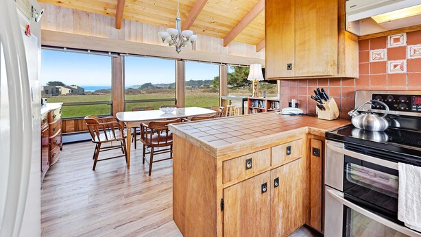 A kitchen with wooden cabinets, a central island, a stove, and a dining area with chairs and a table. - Sea Ranch 3BR distant ocean-view home with hot tub — dogs welcome (Sea Ranch)