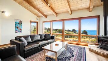 A living room with a black leather sofa, a glass coffee table, a patterned rug, large windows with a view of the sea, and a wood-beamed ceiling.