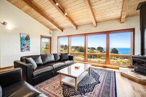 A living room with a black leather sofa, a glass coffee table, a patterned rug, large windows with a view of the sea, and a wood-beamed ceiling. - Ocean-view home with private hot tub near Pebble Beach (Sea Ranch)