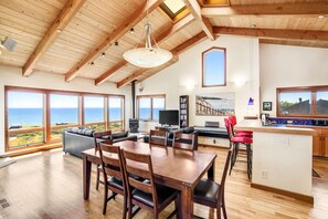 A spacious living area with a wooden ceiling, large windows with ocean views, a dining table, and a kitchen area. - Ocean-view home with private hot tub near Pebble Beach (Sea Ranch)