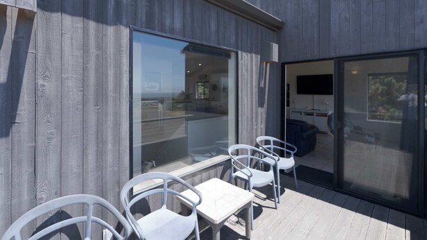 Un edificio moderno con fachada de madera, grandes puertas correderas de vidrio y un patio con sillas y mesa blancos.