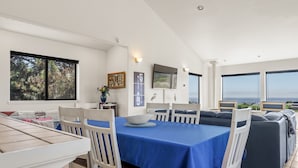 Un comedor moderno con mantel azul, sillas blancas y un ventanal grande con vista al mar.
