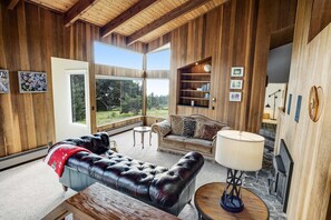 A wooden-paneled room with a leather sofa, a round table, a sofa with cushions, a bookshelf, and a fireplace. - Sea Ranch meadow home with fireplace & panoramic ocean views (Sea Ranch)