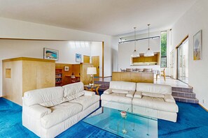A modern living room with a glass coffee table, two white sofas, a kitchen area with a dining table, and a staircase leading to an upper level. - Sea Ranch 2BR Don Jacobs-designed home with hot tub near Shell Beach (Sea Ranch)