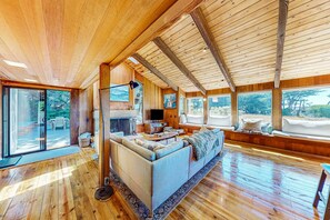 A spacious living area with a large sofa, wooden flooring, and large windows offering views of the outdoors. - Ocean-view home with hot tub & prime location at Shell Beach (Sea Ranch)